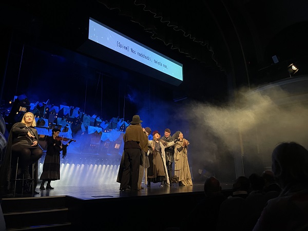 Zdjęcie przedstawia scenę Teatru w trakcie spektaklu, na której znajdują się aktorzy. Po lewej stronie siedzi Pani tłumacz języka migowego, a nad sceną wyświetlone są napisy.