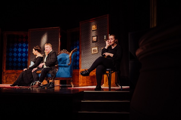 Na zdjęciu widoczny jest fragment przedstawienia. Po prawej stronie sceny siedzi tłumaczka języka migowego. Za nią, w tle sceny widoczni są aktorzy siedzący na zabytkowej sofie.