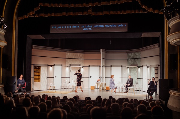 Zdjęcie przedstawia widok całej sceny podczas spektaklu. Po obu stronach siedzą tłumaczki języka migowego, po jednej dla każdego "pokoju hotelowego". Na środku sceny aktorzy odgrywają przedstawienie. Nad sceną znajduje się ekran wyświetlający napisy.