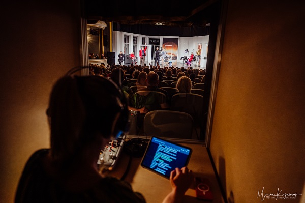 Na zdjęciu przedstawiony jest widok z budki lektora audiodeskrypcji. Aktorka Teatru - Małgorzata Osiej - Gadzina siedzi przy konsoli sterujące zdjęciem. Przed nią znajduje się tablet z wyświetlonym skryptem.