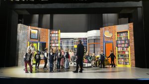 Grupa dzieci jest na scenie teatralnej. Scenografia przedstawia pokój dzienny. 