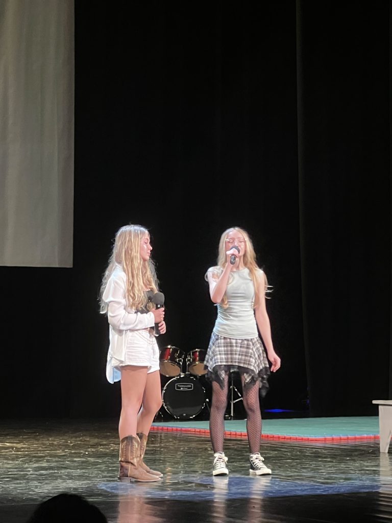 Dwie dziewczynki w blond włosach stoją i śpiewają na scenie.