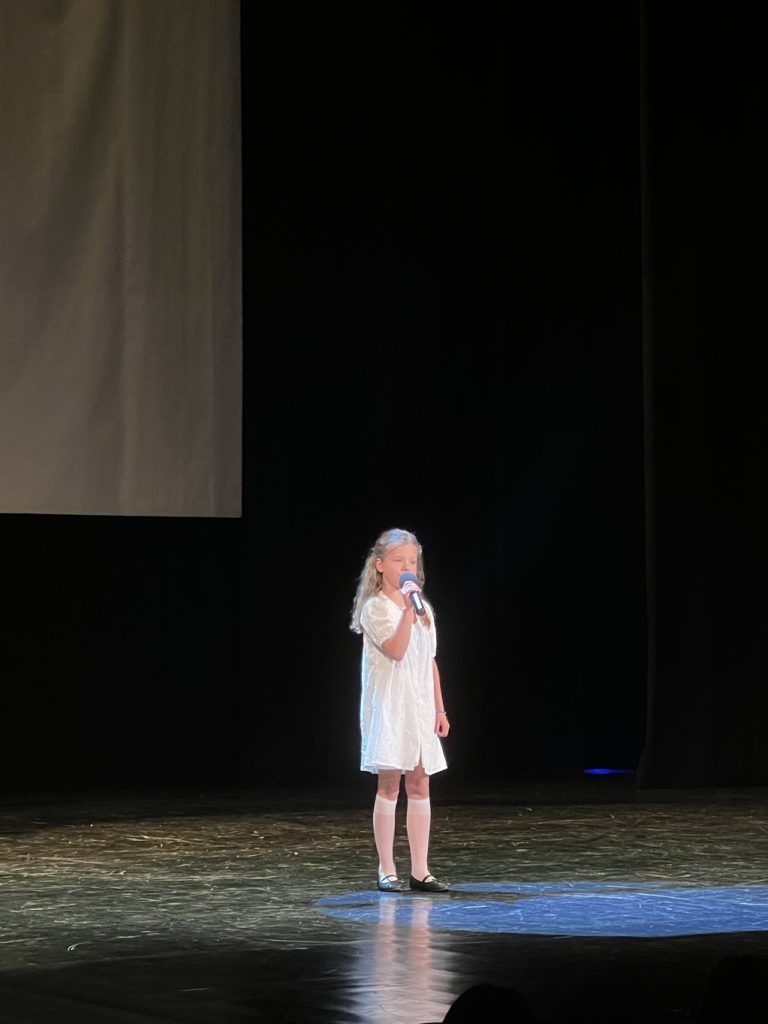 Dziewczynka w blond włosach i białej sukience śpiewa na scenie.