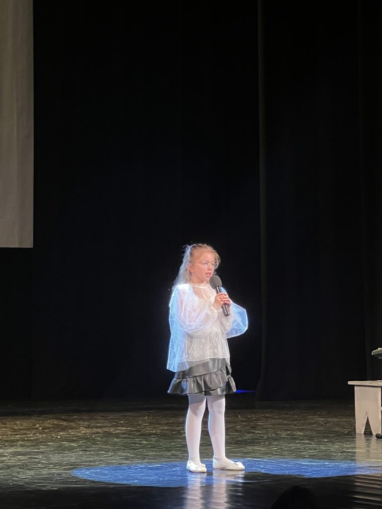 Dziewczynka w blond włosach, białej koszulce i spódniczce śpiewa.