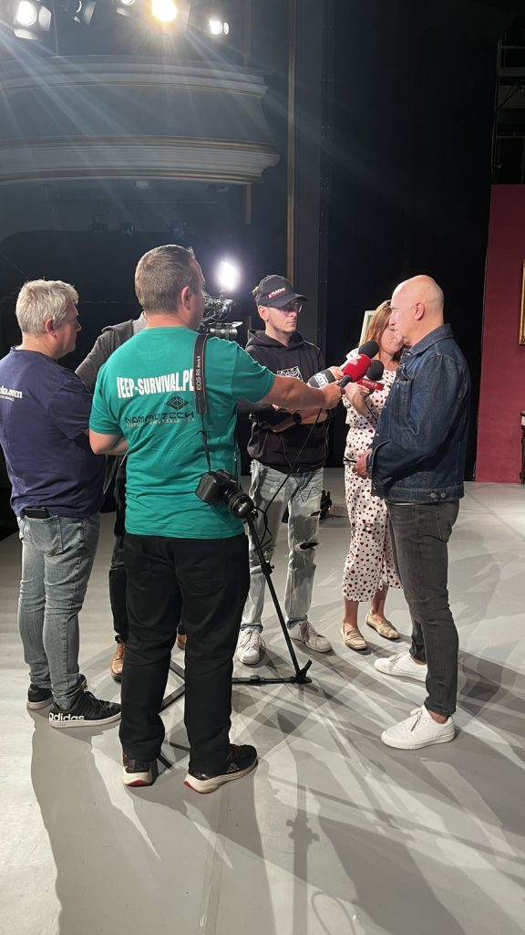 Ludzie z prasy stoją za kamerą i trzymają mikrofony. Przed kamerą stoi mężczyzna.