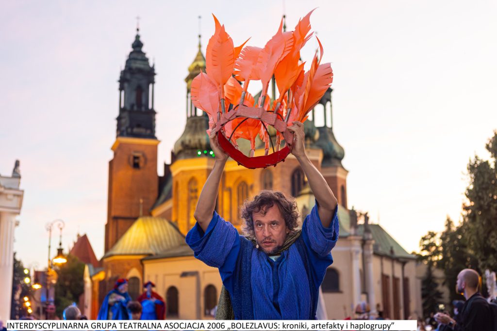 Mężczyzna w niebieskim stroju unosi nad głową koronę z pomarańczowych piór. W tle kościół.
