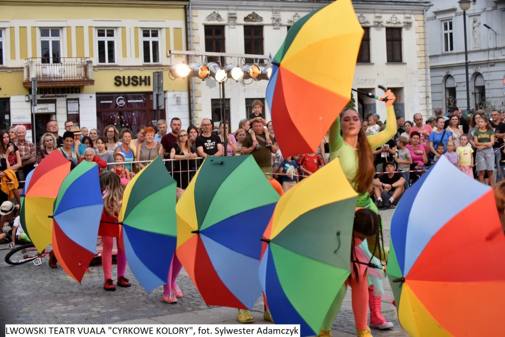 Sześć osób trzyma kolorowe parasolki dołem. Wszyscy stoją w szeregu. W tle tłum ludzi.