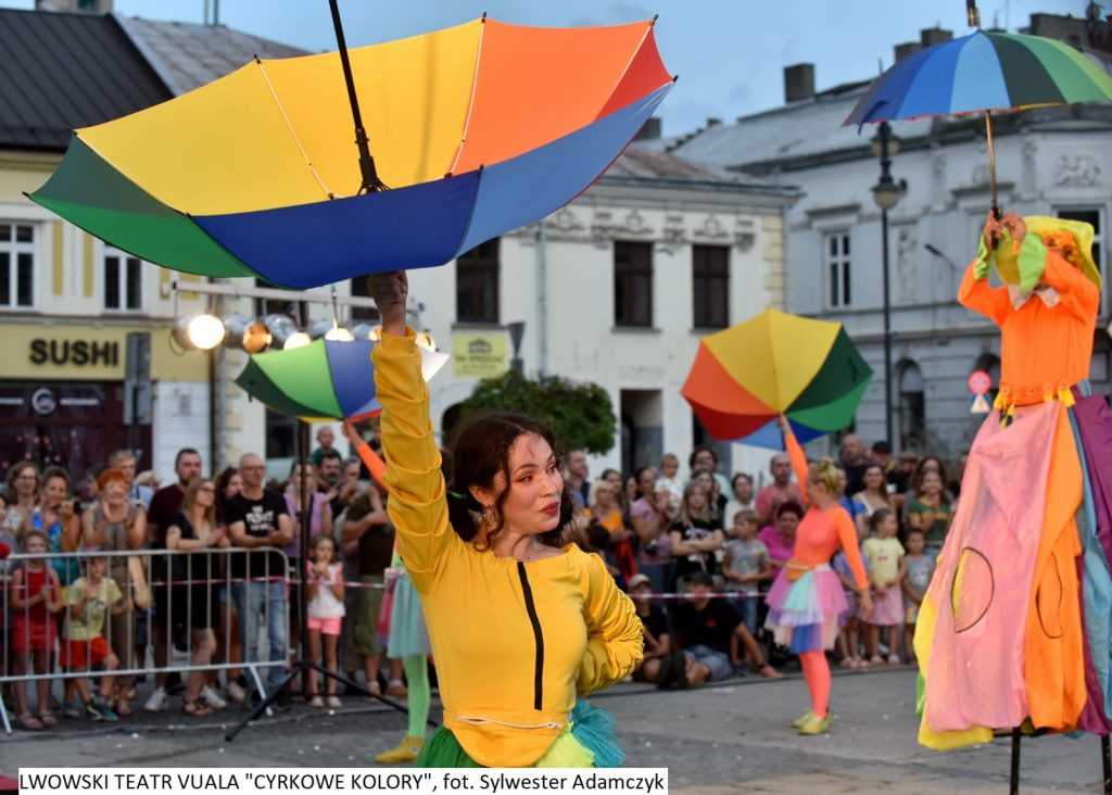 Na pierwszym planie stoi kobieta w żółtej koszulce i trzyma kolorową parasolkę nad głową do góry nogami. W tle dwie inne dziewczyny robią to samo. Po prawej stronie stoi osoba na szczudłach ubrana kolorowo. W oddali tłum ludzi.