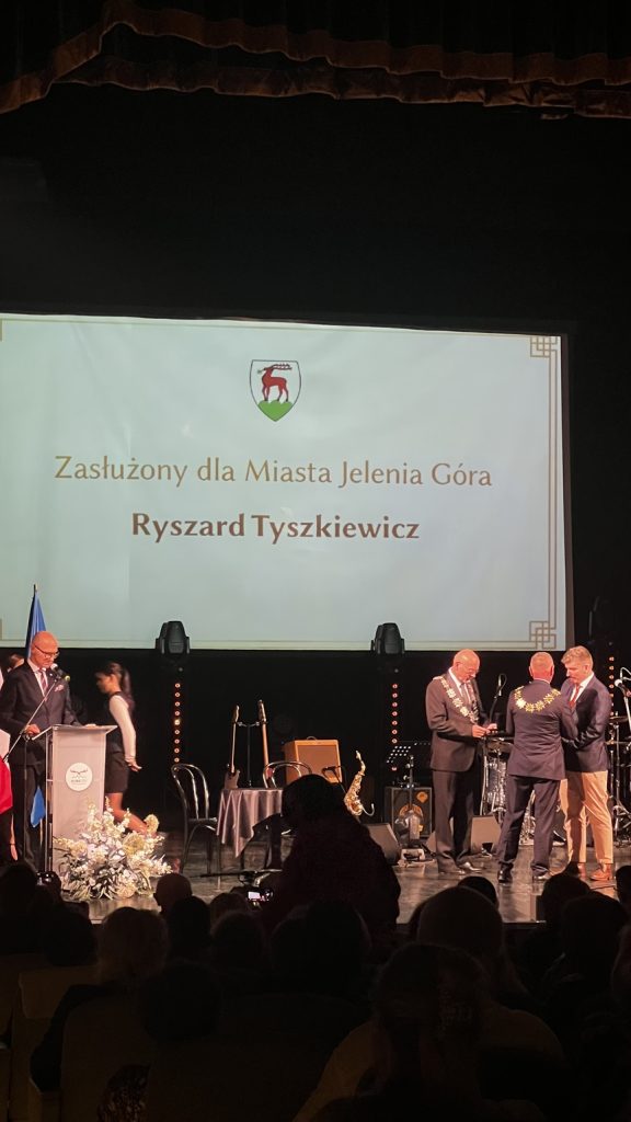 Na scenie czterech mężczyzn ubranych w garniturach. Jeden za mównicą a dwóch wręcza nagrodę czwartemu.