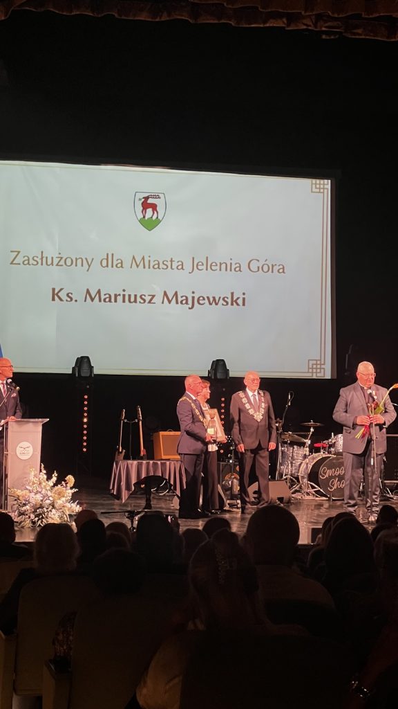 Czterech mężczyzn na scenie i jedna kobieta. Jeden mężczyzna stoi za mównicą, drugi stoi przy mikrofonie, a dwóch mężczyzn i kobieta stoją razem trochę dalej na lewo.