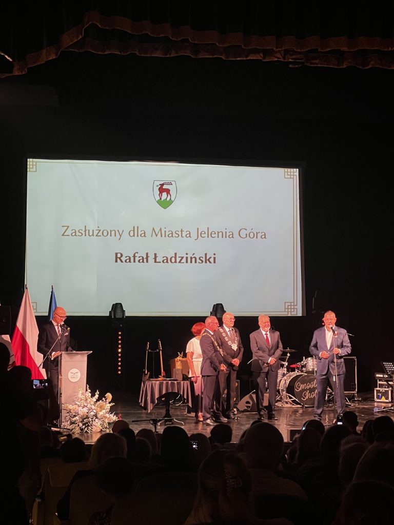 Pięciu mężczyzn w garniturach na scenie i jedna kobieta.