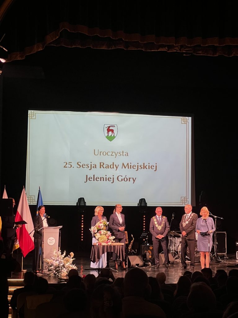 Trzy kobiety, w tym dwie przy mikrofonach oraz trzech mężczyzn. Wszyscy stoją na scenie.