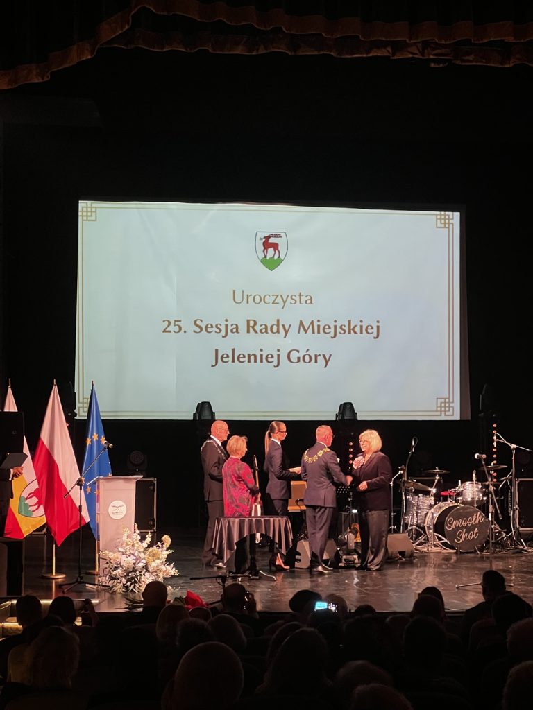 Dwóch mężczyzn w garniturach i jedna kobieta wręczają upominki drugiej kobiecie.