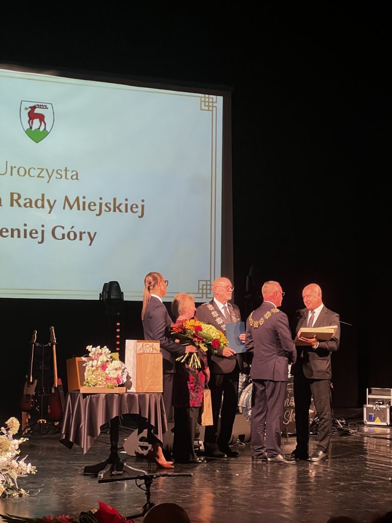 Dwóch mężczyzn w garniturach wręczają nagrodę trzeciemu mężczyźnie.