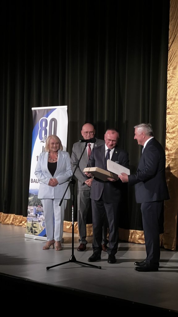 Trzej mężczyźni w garniturach i kobieta w jasno niebieskim garniturze. Wszyscy stoją na scenie.