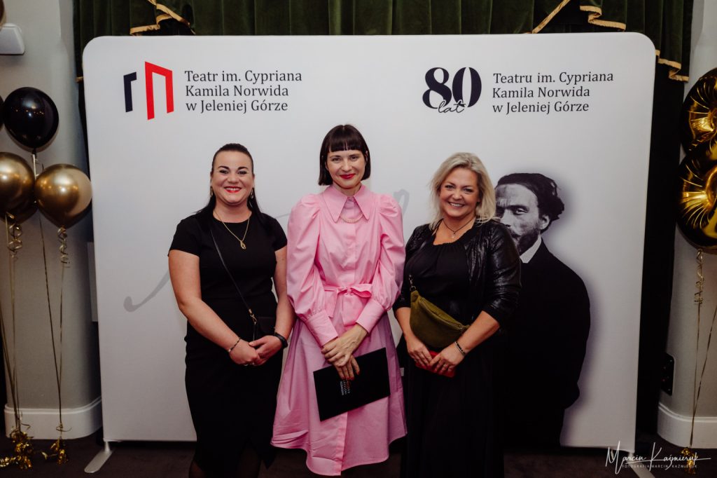 Trzy kobiety stoją elegancko ubrane przed ścianką do zdjęć. Jedna jest ubrana na różowo, reszta na czarno.