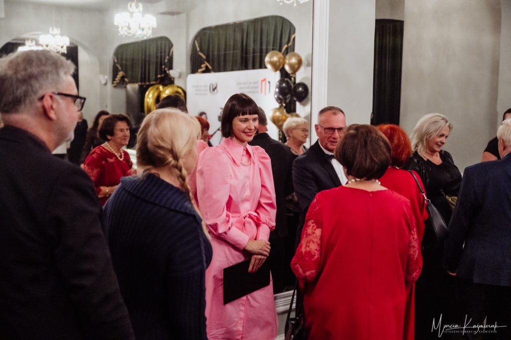 Kobieta ubrana na różowo otoczona przez grupę elegancko ubranych osób.