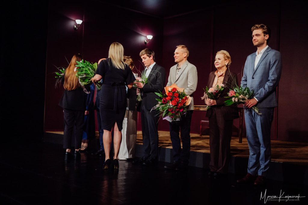 Kobieta elegancko ubrana rozdająca kwiaty osobom na scenie.