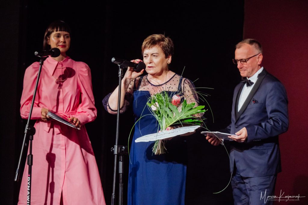 Dwie kobiety i mężczyzna stojący na scenie, jedna kobieta mówi do mikrofonu.