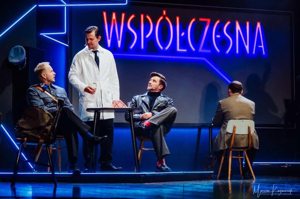 Trzech mężczyzn siedząca na krzesłach zwrócona do mężczyzny ubranego w kitel.