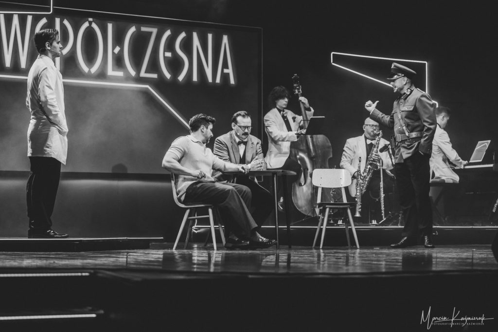 Dwóch mężczyzn siedząca przy stoliku, przed mężczyznami grającymi na instrumentach.