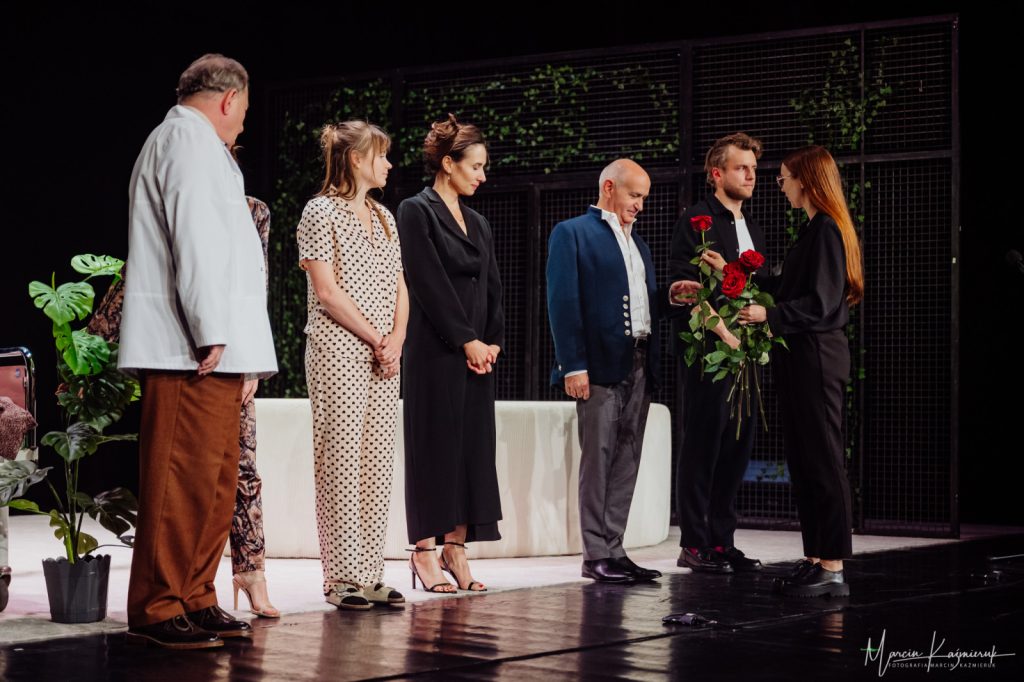 Kobieta ubrana na czarno rozdająca kwiaty osobom stojącym na scenie.