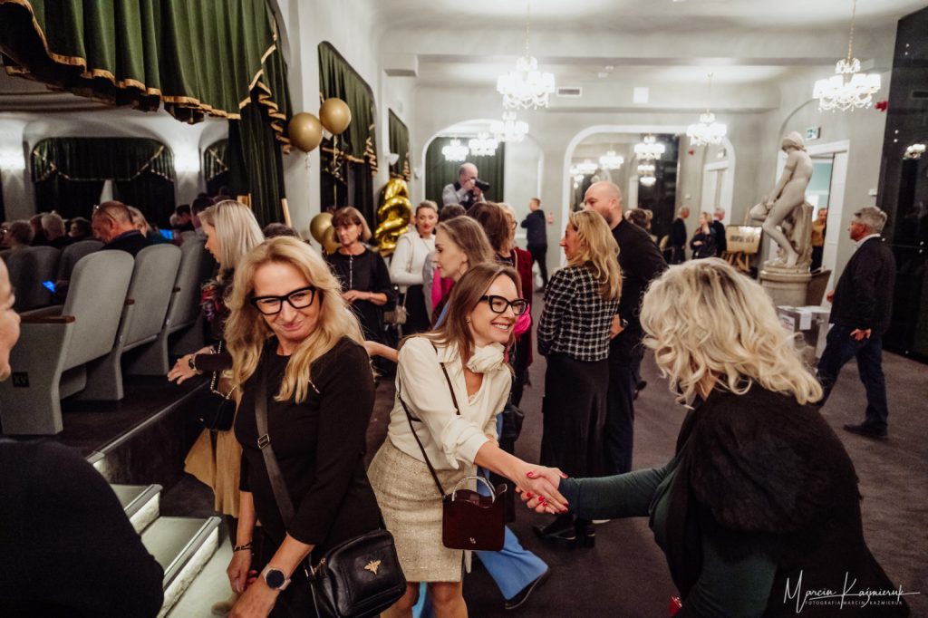 Grupa osób czekająca pod salą teatralną. Dwie kobiety witają się, podając sobie rękę.