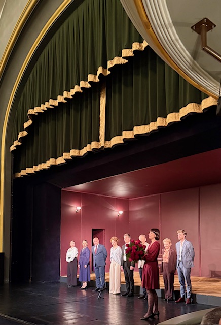 Scena w teatrze a na niej grupa ludzi elegancko ubranych.