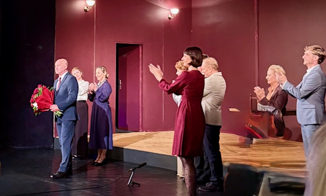 Ludzie na scenie stoją i klaszczą na scenie. Mężczyzna ubrany w garnitur stoi z kwiatami.