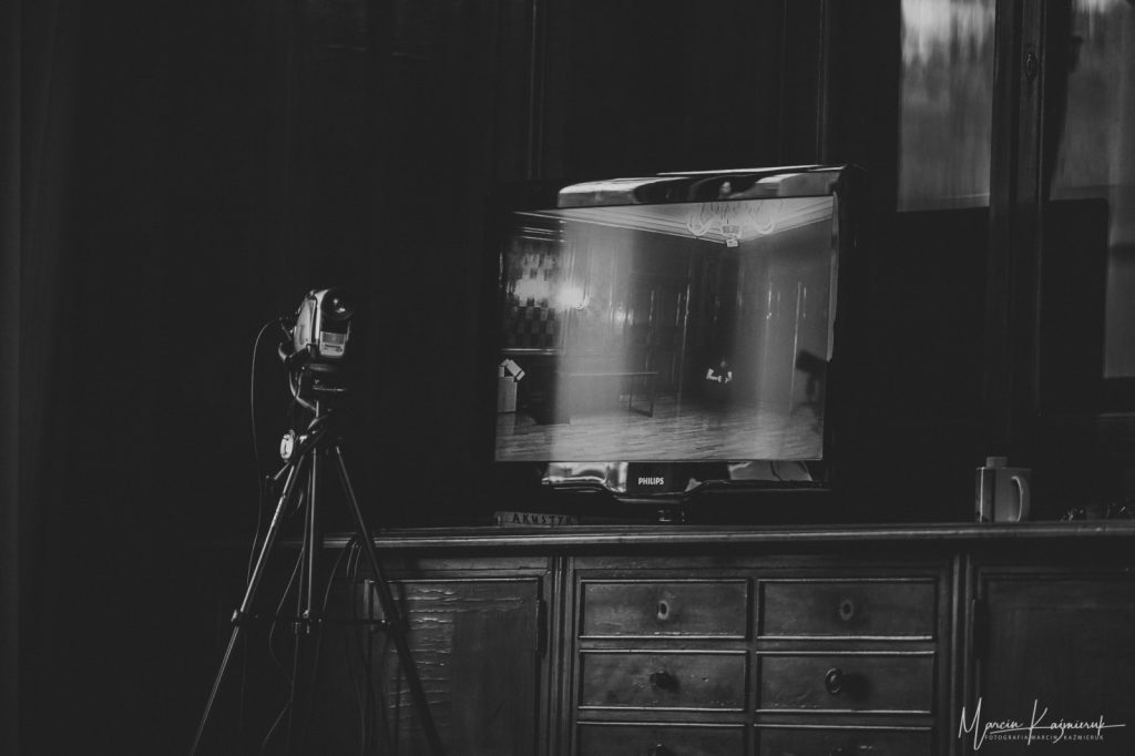 Na komodzie stoi telewizor z płaskim ekranem. Przed nim stoi na statywie kamera.