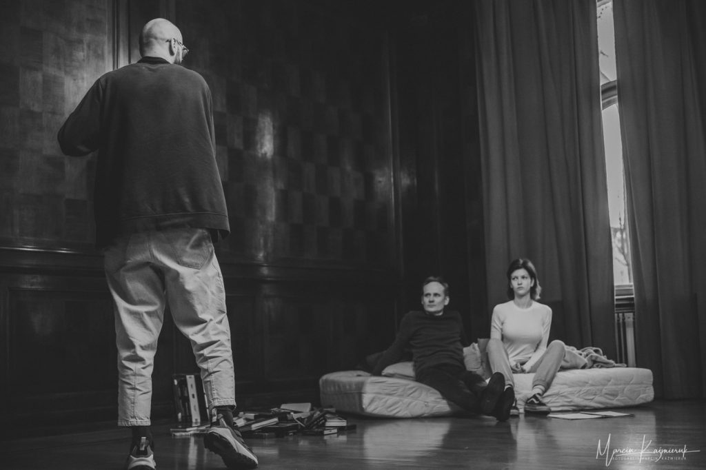 Na materacu w rogu pomieszczenia siedzi mężczyzna po lewej i dziewczyna po prawej. Przed nimi stoi łysy mężczyzna tyłem do fotografa.