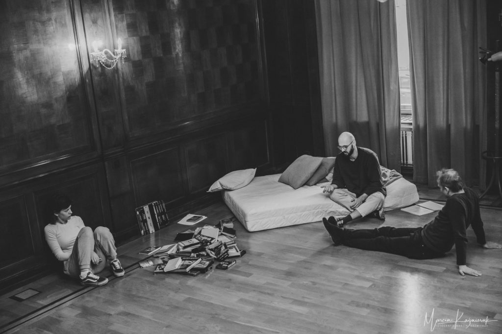 W rogu pomieszczenia na materacu siedzi łysy mężczyzna w okularach. Po jego lewej na podłodze siedzi drugi mężczyzna z wyprostowanymi nogami. Pod ścianą siedzi dziewczyna.