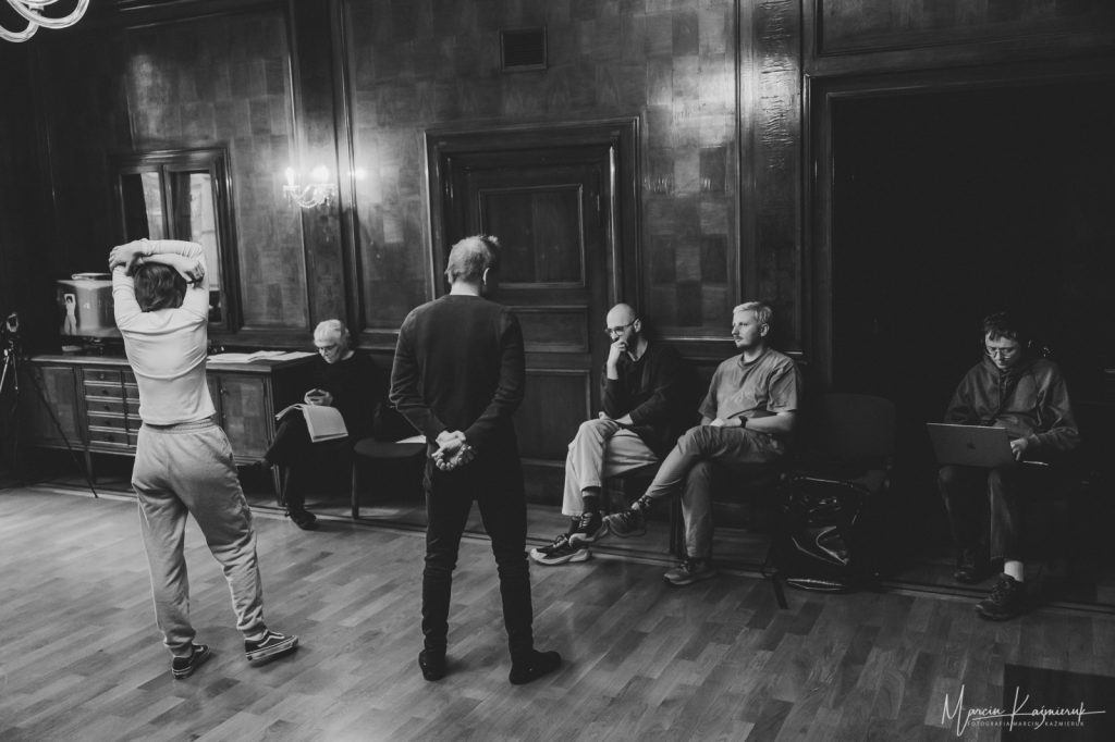 Cztery osoby siedzą pod ścianą. Od lewej kobieta, kolejno potem mężczyźni. Przed nimi stoją przodem dziewczyna i mężczyzna.