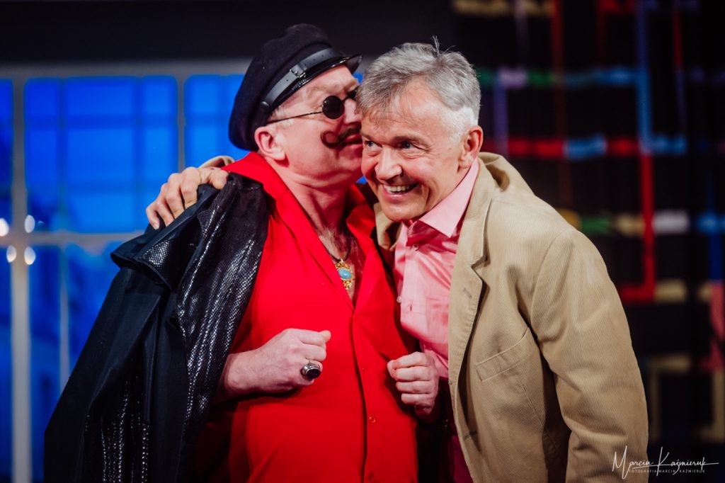 Dwóch mężczyzn się obejmuje. Po lewej mężczyzna ma wąsy, czarne okrągłe okulary, czarną czapkę, czarny płaszcz i czerwoną koszulę. Po prawej stronie mężczyzna sie uśmiecha, bo ten po lewej mówi mu coś na ucho. Mężczyzna po prawej ma brązowa marynarkę i różową koszulę.