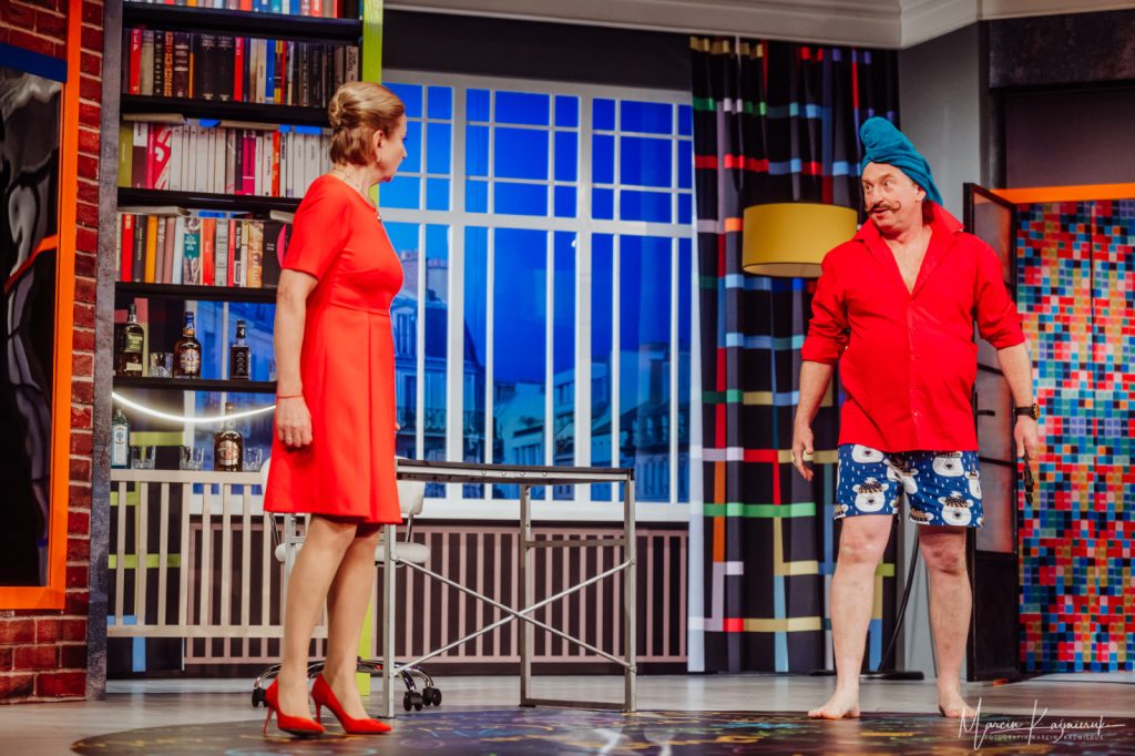 Przestrzeń to pokój z regałami na książki, na środku znajduje się okno. Po lewej stronie stoi kobieta w czerwonej sukience i czerwonych obcasach. Patrzy na mężczyznę stojącego po prawej stronie, który ma na sobie niebieskie spodenki z misiami, czerwoną koszulę i turban z ręcznika na głowie.