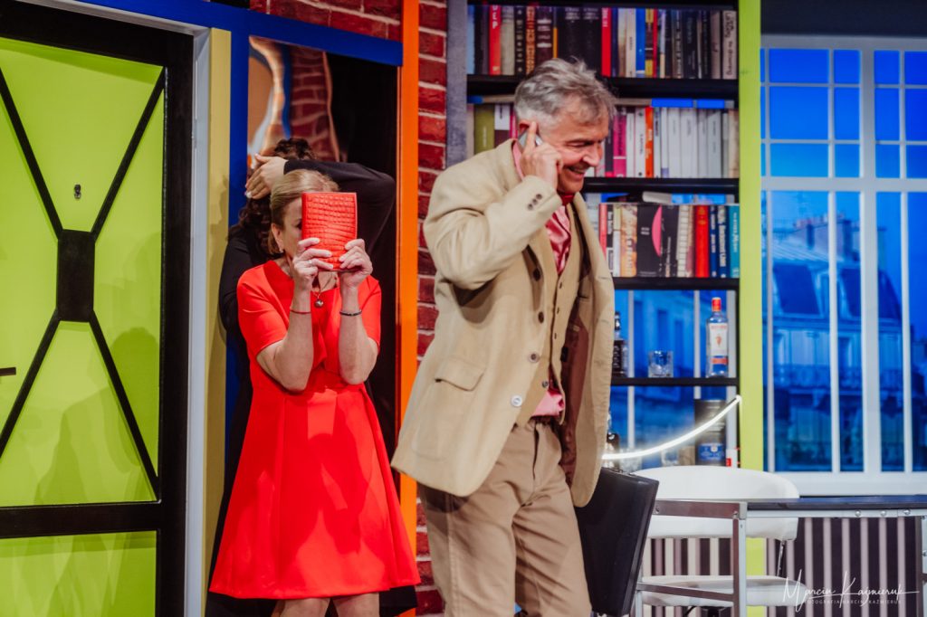 Na pierwszym planie stoi bokiem mężczyzna rozmawiający przez telefon. Ma na sobie brązowy garnitur i różową koszulę. W lewej ręce trzyma teczkę. Za nim stoi kobieta w czerwonej sukience, zakrywa twarz czerwoną torebką.