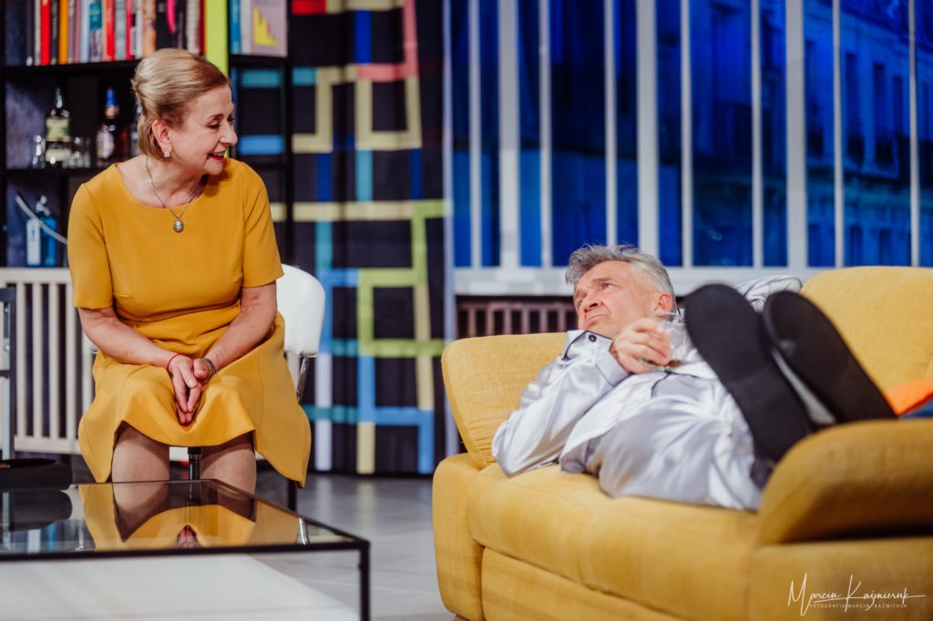 Po lewej stronie zdjęcia stoi szklany stolik. Po prawej stronie stoi żółta sofa, na której leży mężczyzna w srebrnej piżamie z długimi rękawami. Po jego prawej na krześle siedzi kobieta w żółtej sukience.