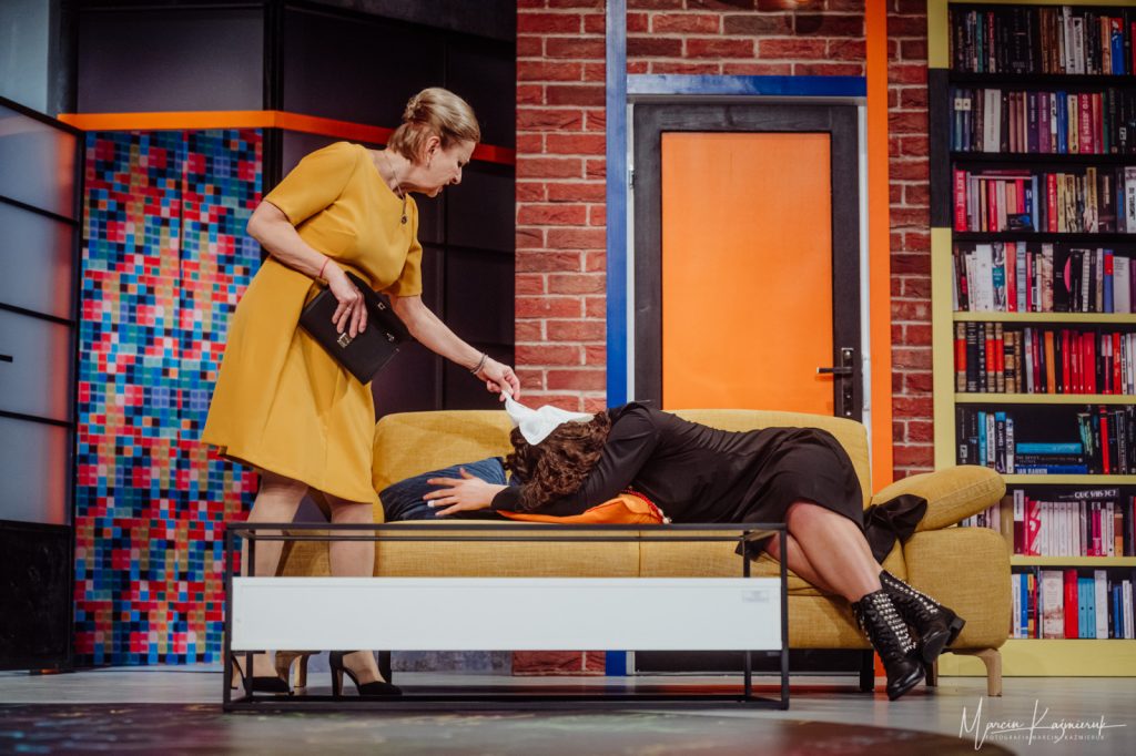 Na pierwszym planie stoi biały stolik kawowy. Na drugim planie stoi żółta sofa. Na niej leży kobieta ubrana w czarne kozaki i czarną sukienkę. Ukrywa twarz w poduszce. Nad nią stoi druga kobieta ubrana w żółtą sukienkę.