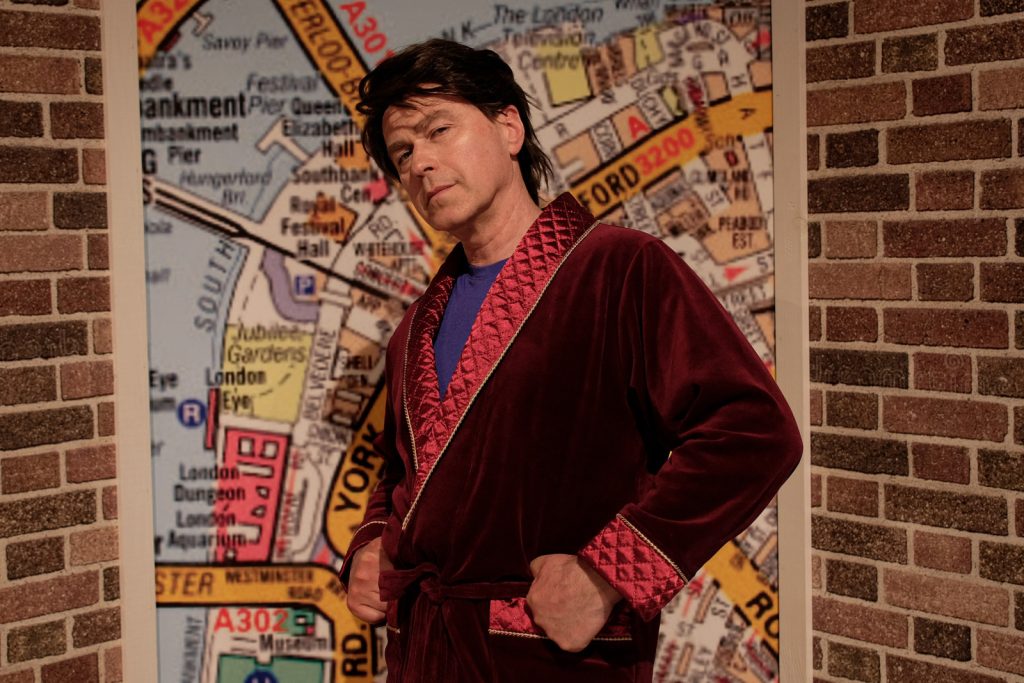 Mężczyzna w ciemnych krótkich włosach stoi w bordowym szlafroku bokiem. Ma schowane ręce w kieszeni.