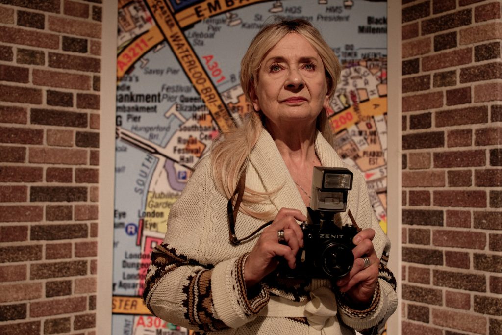 Kobieta w blond włosach stoi i w rękach trzyma aparat. Patrzy w dal. Ma na sobie beżowy sweter.