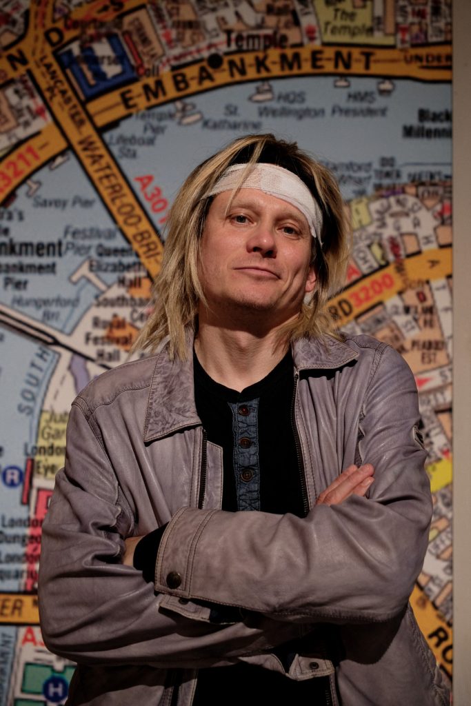 Mężczyzna w blond włosach i bandażu na głowie stoi na wprost. Nosi szarą koszulę. Ręce ma założone na krzyż na piersi.