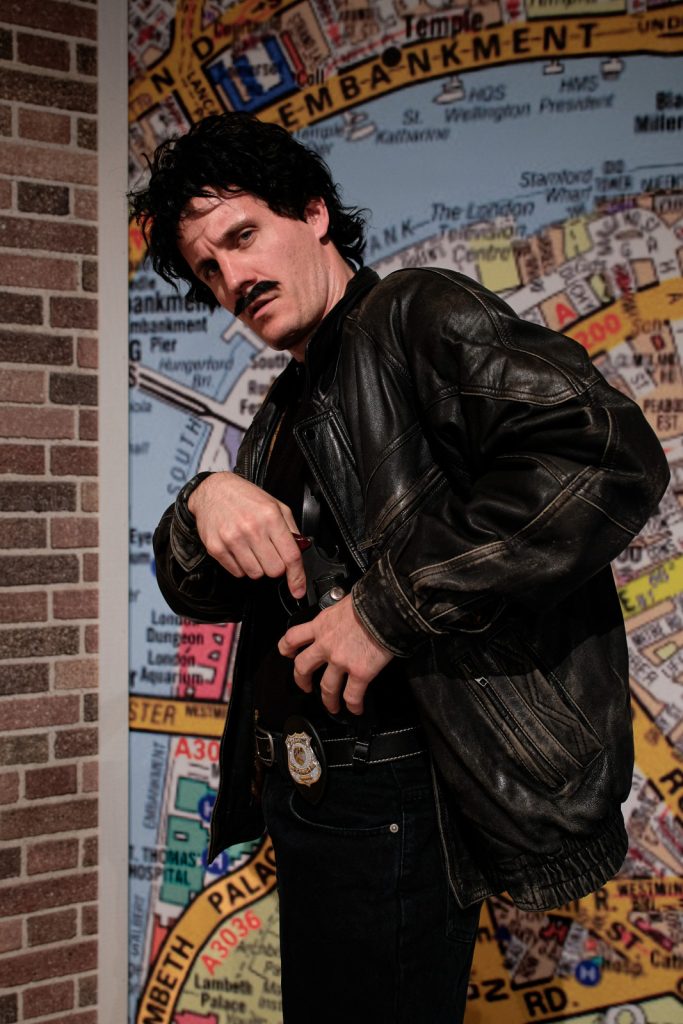 Mężczyzna w czarnych krótkich włosach oraz wąsach. Chowa pistolet do spodni. Jest ubrany na czarno.