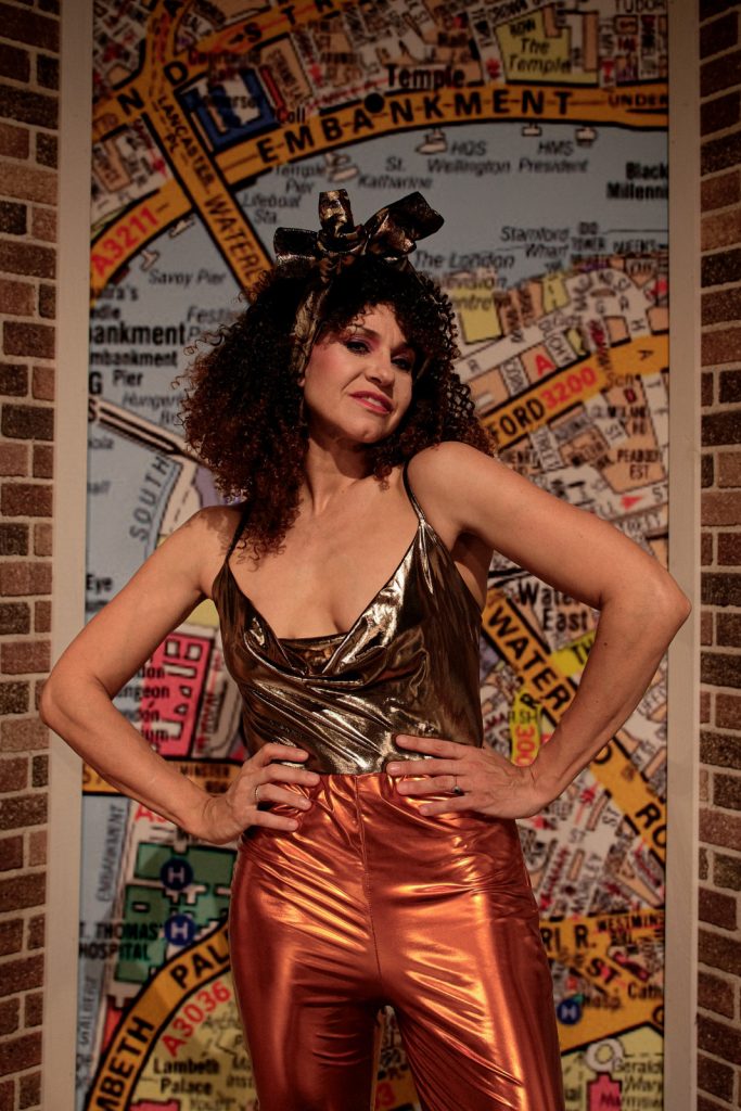 Kobieta w kręconych brązowych włosach stoi z założonymi rękoma na biodra. Spodnie ma pomarańczowe i koszulkę na ramiączkach.