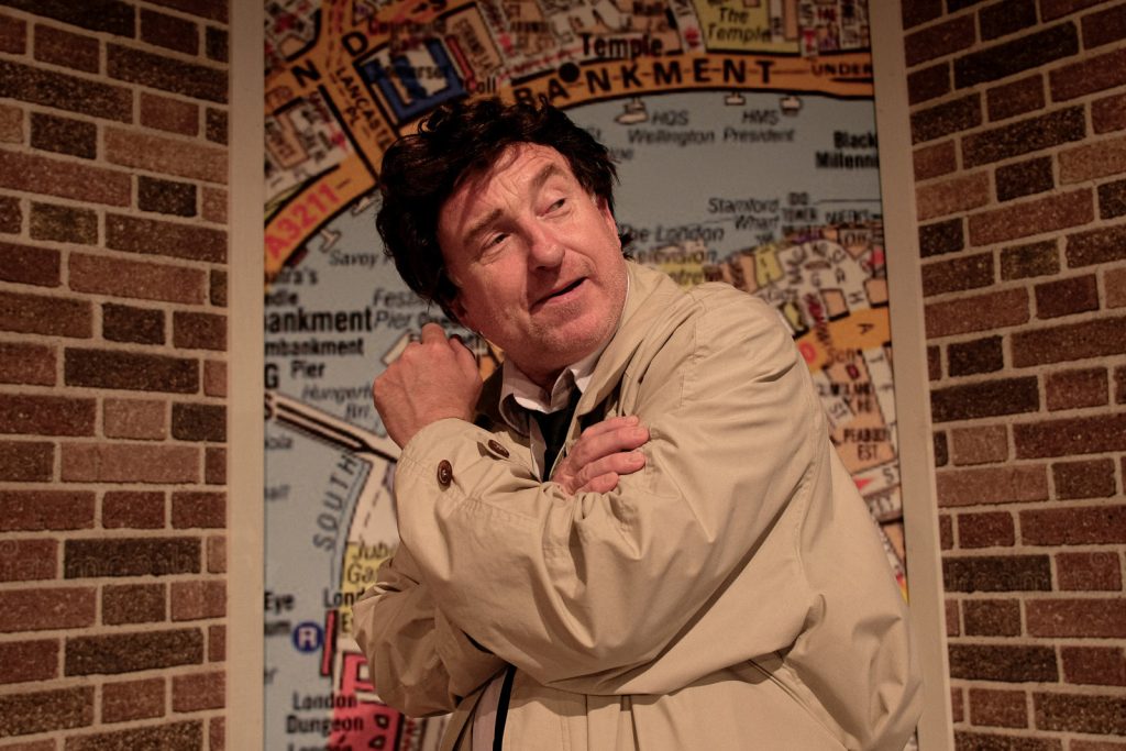 Mężczyzna w brązowych włosach w beżowym płaszczu stoi bokiem. Patrzy się przez lewe ramie.