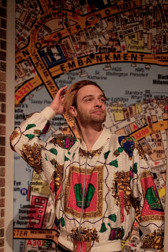 Młody mężczyzna w blond włosach stoi i patrzy się w dal. Prawą rękę trzyma na głowie. Ma na sobie wzorzystą bluzę.