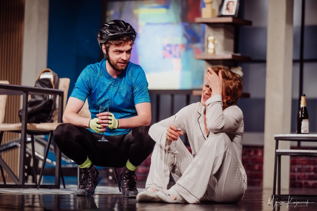 Kobieta siedzi na ziemi ze zgętymi nogami i kieliszkiem w ręki. Opiera głowę o rękę. Koło niej kuca mężczyzna w kasku i niebieskiej koszulce.